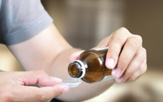 Das Bild zeigt eine Nahaufnahme einer Person, die Medizin aus einer braunen Flasche in einen Messlöffel gießt. Die Person trägt ein hellgraues T-Shirt. Der Hintergrund ist unscharf, aber es scheint sich um einen Innenraum zu handeln. Das Bild vermittelt einen Eindruck von Krankheit oder Unwohlsein und der Notwendigkeit, Medikamente einzunehmen. Die Handlung ist einfach, aber die Konzentration auf die Hände und die Flasche betont die Intimität des Aktes der Selbstmedikation.