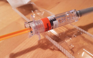 Das Bild zeigt eine einzelne, gebrauchsfertige Spritze in einer durchsichtigen Plastikverpackung auf einer Holzoberfläche. Die Spritze ist aus transparentem Kunststoff gefertigt und enthält eine klare Flüssigkeit. Ein orangefarbener Kolben ist sichtbar. Ein orangefarbenes Etikett um den Spritzenkörper weist auf den Inhalt hin. Die Verpackung deutet darauf hin, dass es sich um ein medizinisches Produkt handelt, das für den einmaligen Gebrauch bestimmt ist. Das Bild vermittelt einen Eindruck von Sauberkeit und Sterilität.