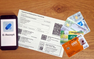 Das Bild zeigt ein Smartphone mit der E-Rezept-App, einen Ausdruck eines elektronischen Rezepts und mehrere deutsche Gesundheitskarten. Es illustriert die digitale Transformation im deutschen Gesundheitswesen, mit dem Fokus auf die zunehmende Nutzung von E-Rezepten und digitalen Gesundheitskarten. Das Bild vermittelt einen Eindruck von Effizienz und Modernisierung im Gesundheitssektor, indem es die papierlose Abwicklung von Rezepten und die vereinfachte Identifizierung des Patienten durch digitale Karten zeigt. Die verschiedenen Gesundheitskarten deuten auf die Vielfalt der Krankenkassen in Deutschland hin. Insgesamt präsentiert das Bild eine moderne und effiziente Methode der Gesundheitsversorgung.