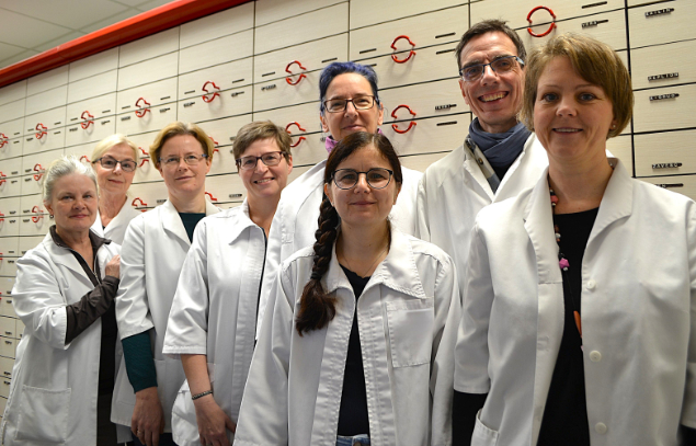 Das Foto zeigt fünf Apothekerinnen in weißen Kitteln, die in einer Apotheke zusammenarbeiten. Sie scheinen sich über ein Buch oder eine Broschüre zu beugen, die auf einem Tresen liegt. Die Atmosphäre ist freundlich und kollegial. Die Frauen wirken konzentriert und professionell, gleichzeitig aber auch entspannt und zugewandt zueinander. Das Bild strahlt Kompetenz und Teamwork aus und vermittelt ein Gefühl von Zuverlässigkeit und Sorgfalt, typisch für den Apothekenberuf. Die Anordnung der Frauen und die Beleuchtung des Bildes unterstützen diese positive und harmonische Stimmung.