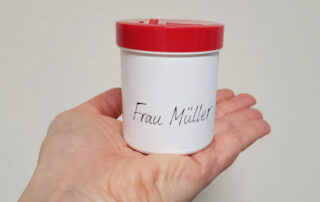 Das Bild zeigt eine Hand, die ein kleines, weißes Gefäß mit einem roten Deckel hält. Auf dem Gefäß steht mit schwarzer Schrift „Frau Müller“. Das Bild ist einfach und unauffällig, könnte aber verschiedene Bedeutungen haben. In dem Gefäß ist die persönliche Rezeptur hergestellt.